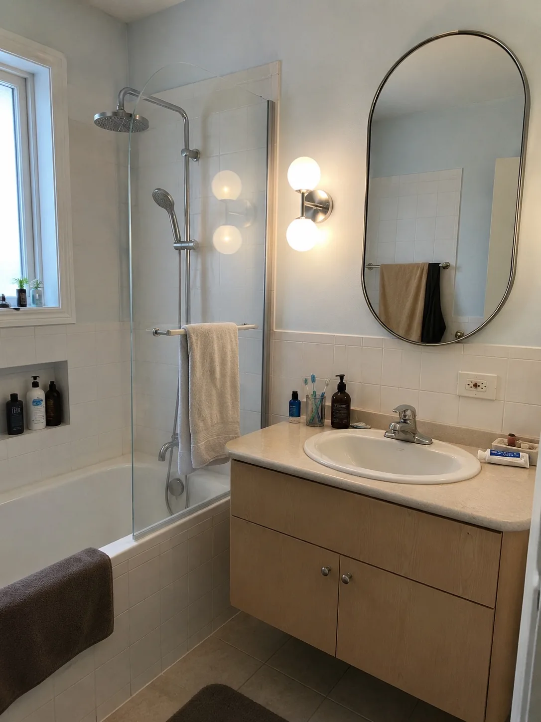 Real bathroom before redesign: white tiled walls, beige laminate vanity, oval mirror, bathtub with a glass shower screen