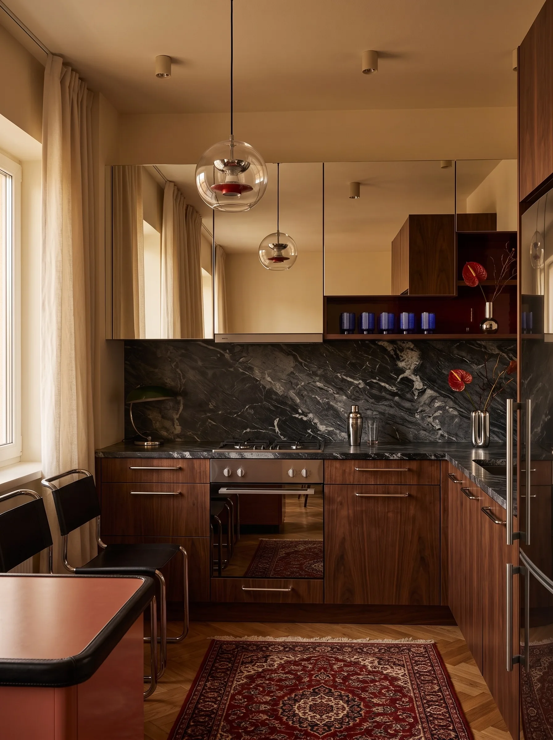 Moody luxe kitchen with walnut cabinets, a black marble splashback and worktop, mirrored upper cabinet fronts, chrome fittings, cantilever chairs and a Persian rug
