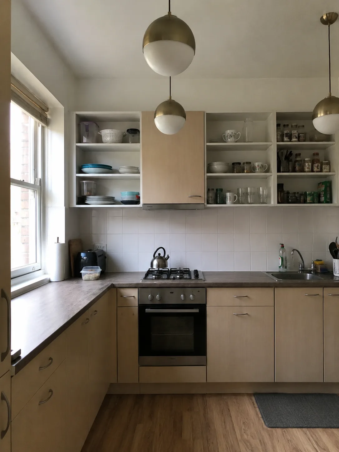 Real kitchen before redesign: L-shaped birch laminate units with doorless upper shelves, grey worktop, white square tile splashback and three brass globe pendants