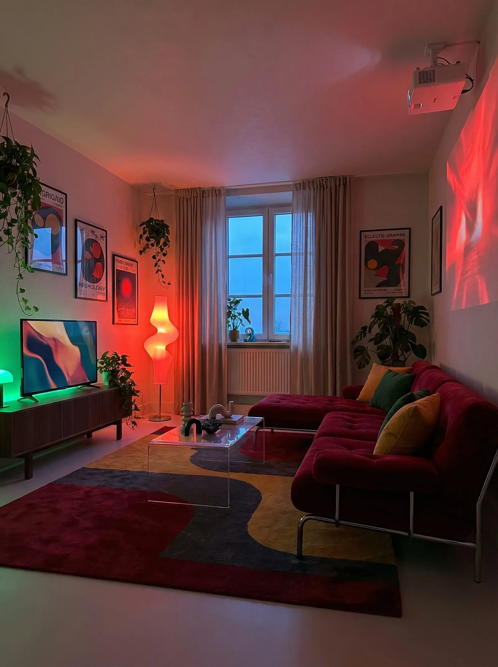 A dusk scene with the TV back on the low walnut console on the left, green bias lighting behind the screen, a deep burgundy velvet tufted sectional with chrome legs, a clear acrylic waterfall coffee table, a wavy burgundy and mustard rug, graphic posters on the walls, a sculptural glowing floor lamp, and a projector mounted on the ceiling.