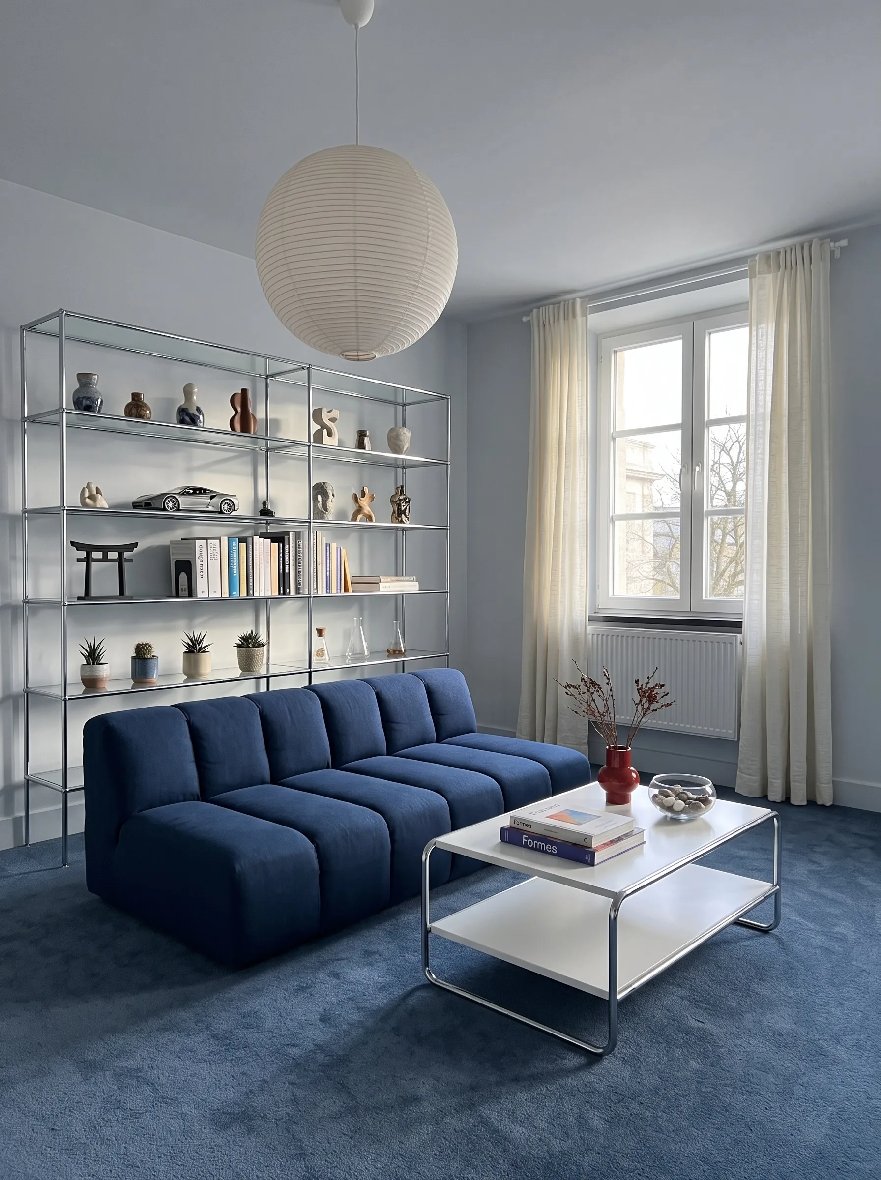 Colour block living room with pale blue walls, a cobalt blue carpet, a navy channel-tufted sofa, a chrome etagere of sculptures and ceramics, a white and chrome coffee table and a paper globe pendant